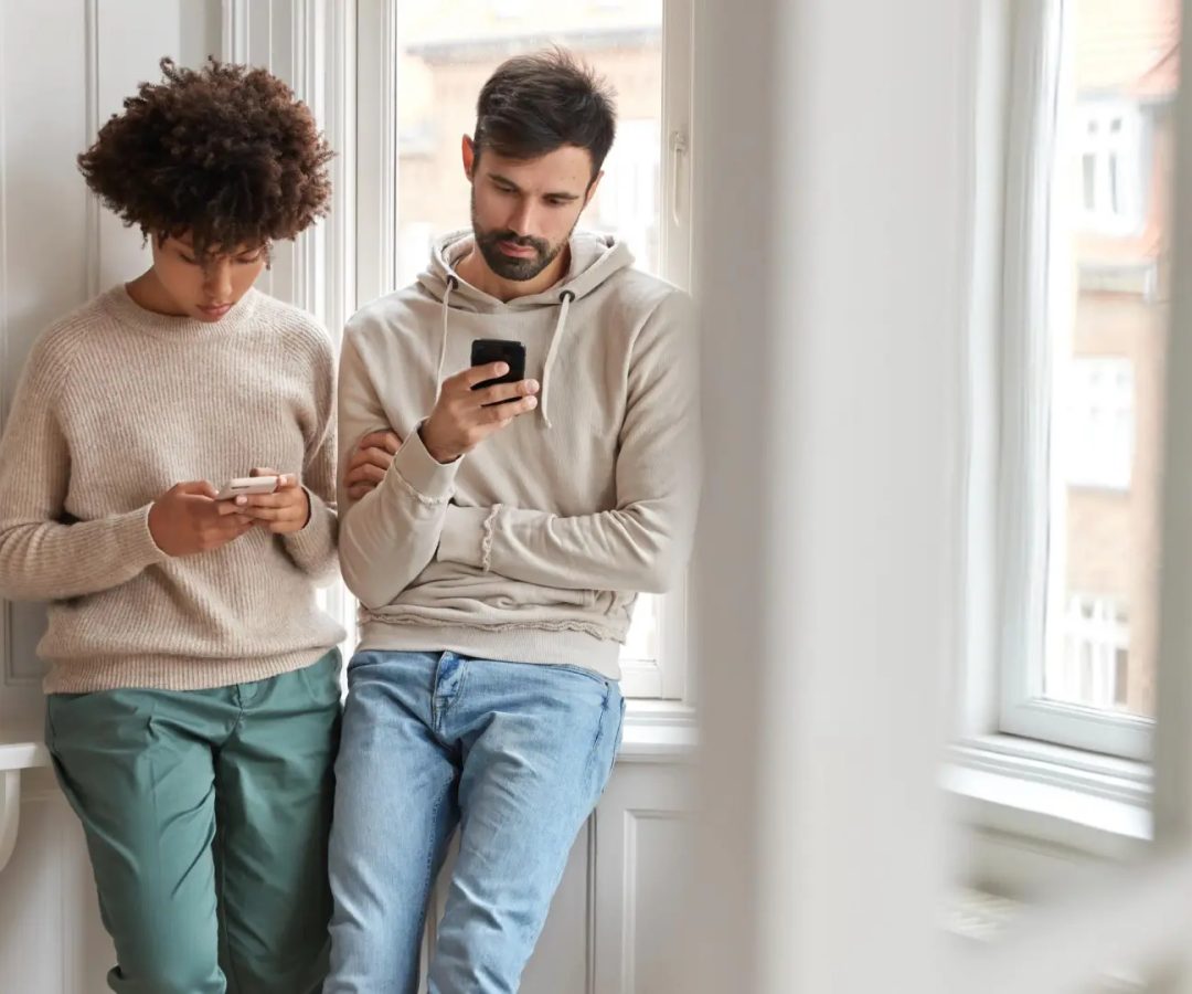 Dos jóvenes, de pie junto a una ventana, concentrados en sus teléfonos móviles.Freepik
