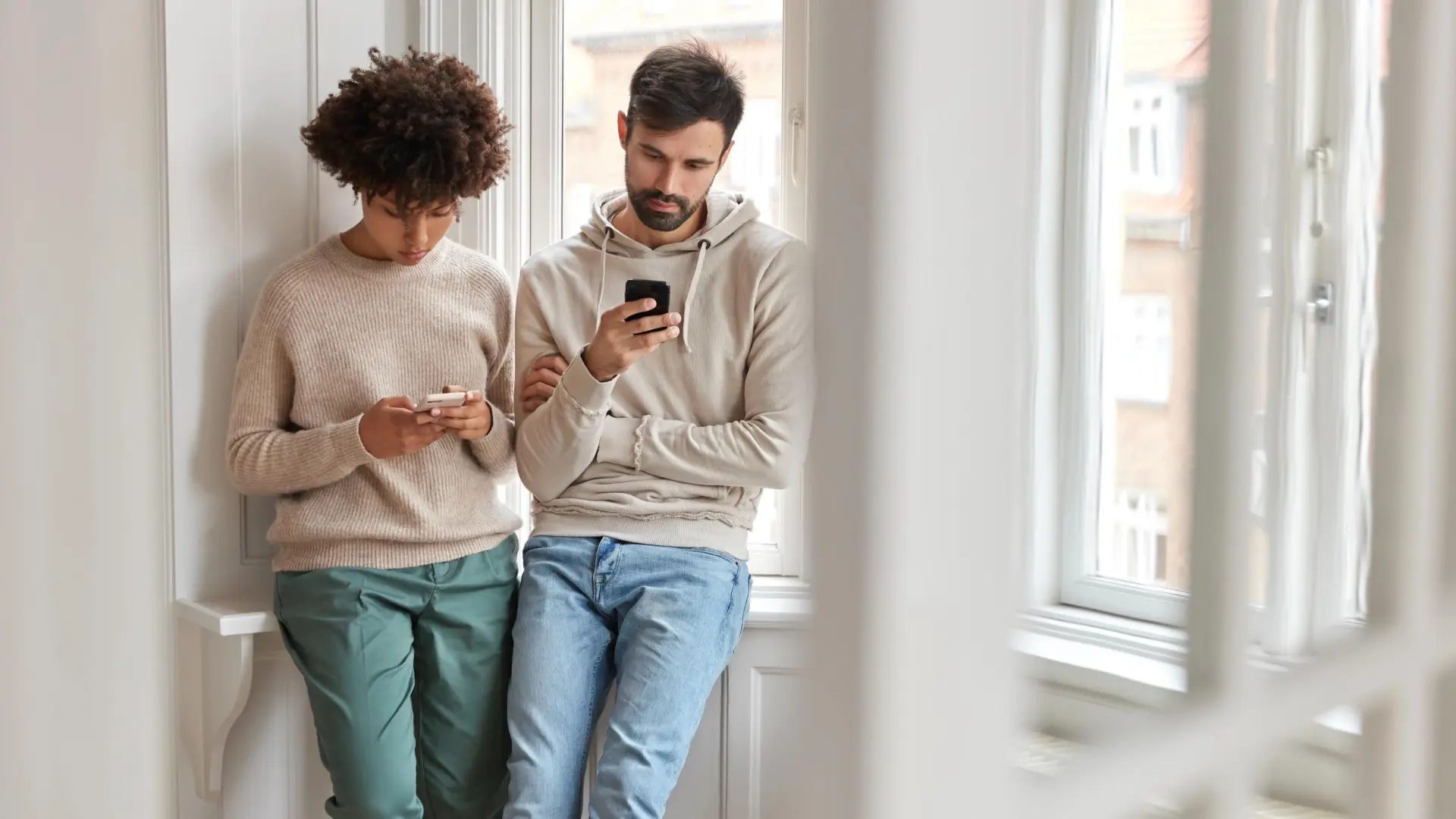 Dos jóvenes, de pie junto a una ventana, concentrados en sus teléfonos móviles.Freepik
